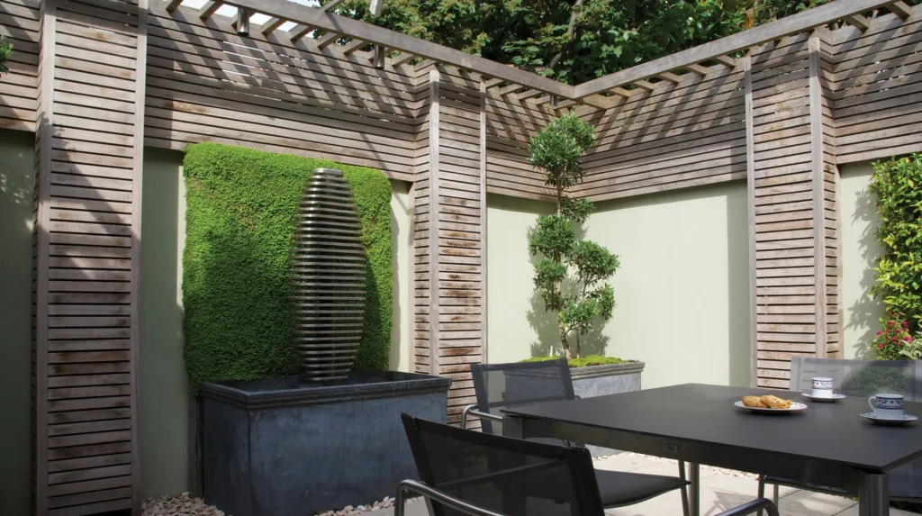 Modern patio with wooden trellis, vertical water feature, plants, and black table set with cups and cookies.