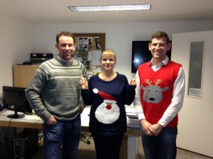 Three people wearing festive sweaters stand together in an office setting.
