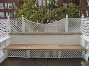 Outdoor wooden bench with white lattice backrest on a patio, with a tree and buildings in the background.