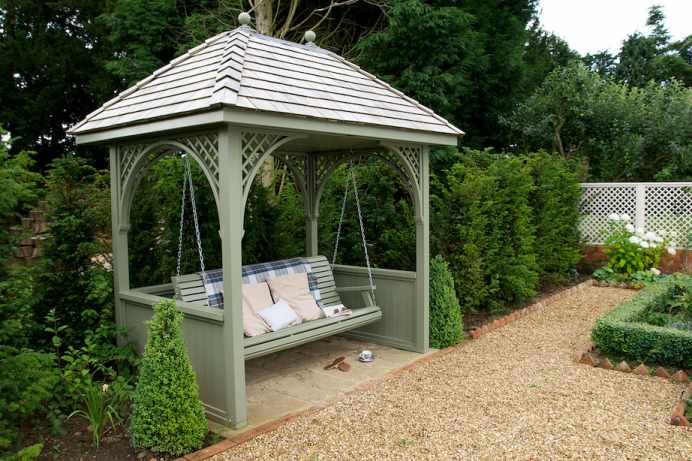Green garden gazebo with a swing bench, cushions, and a plaid blanket, surrounded by greenery and a gravel path.