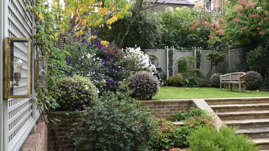 A lush garden with a variety of plants, a wooden bench, and stone steps leading to a lawn, enclosed by a lattice fence.