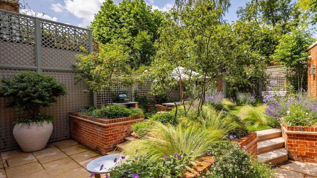 Lush garden with brick planters, tall trees, green shrubs, and purple flowers under a partly cloudy sky.